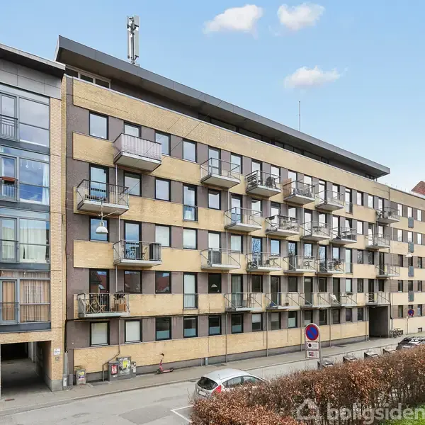 En moderne fem-etagers lejlighedsbygning med altaner på hver etage. Flere biler er parkeret foran bygningen på en stille gade. Omkring er der buske og træer.