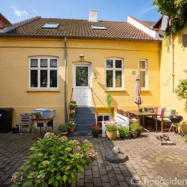 Gul husfacade med to vinduer og en dør i midten. Foran huset står et bord og stole. Blomster og planter omgiver gårdspladsen med brosten. Luften er klar og himlen blå.