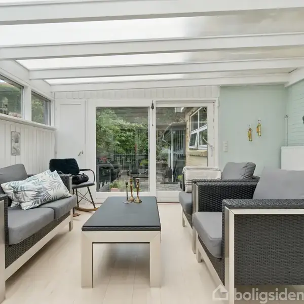 Grå sofaer med puder står omkring et lavt bord. Vinterhave med glasvægge giver udsigt til grønne omgivelser. Rummet er lyst og moderne indrettet.