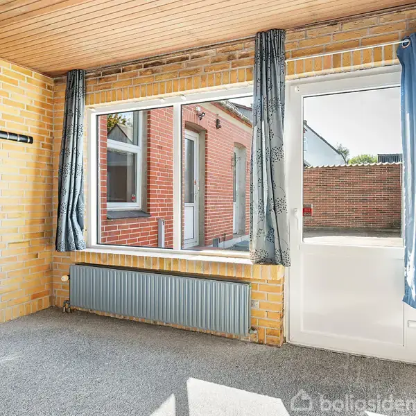 Et værelse med gulv til loft-vindue og dør til gårdhave, omringet af gul murstensvægge og blå gardiner. Udenfor ses en rød murstensvæg og sollys skinner ind.