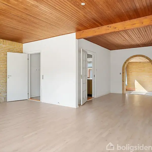 Stue med lyst trægulv, træloft og åbne døre, der fører til andre rum. En bue i baggrunden åbner til et rum med en solbelyst murstensvæg.