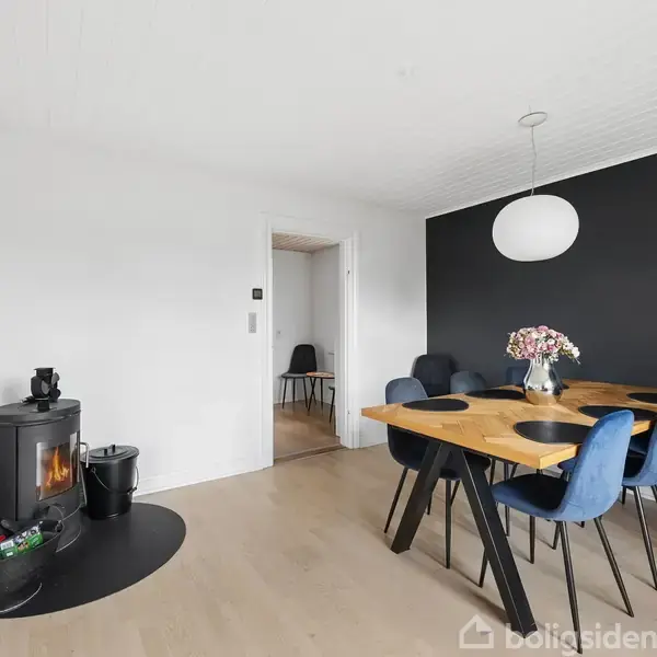 Brændeovn står tæt ved en moderne spisebordsgruppe, hvor seks blå stole omgiver et træbord med blomster i en vase. Rummet har lyse vægge og en mørk accentvæg.