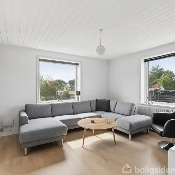 En grå hjørnesofa står i et lyst stuerum med lyse trægulve. Et rundt træbord står foran sofaen, og der er store vinduer med udsigt til en have.