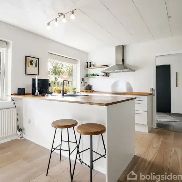 Et lyst, moderne køkken med to høje stole ved en træbordplade. Rummet har store vinduer, hvide vægge og lofter, samt en indbygget emhætte. To potteplanter står på vindueskarmen.
