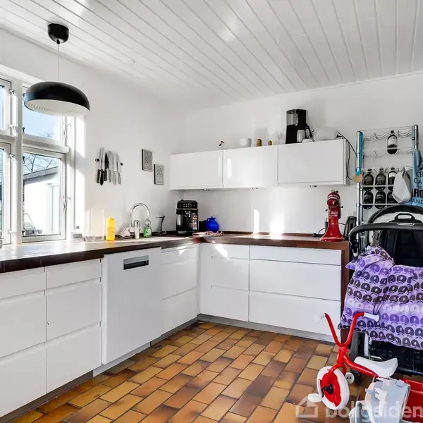 Køkken med hvide skabe og trægulv, en kaffemaskine og juicepresser på bordet. En sort lampe hænger over vasken. En klapvogn og en rød legetøjsmotorcykel står i hjørnet.