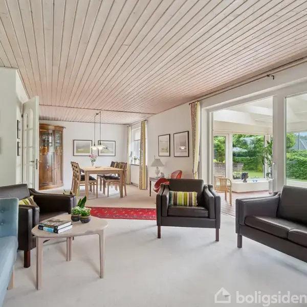 Stue indrettet med blå og sorte sofaer, placeret omkring et sofabord med planter og bøger; stor vinduesfront åbner mod terrasse med udsigt til have.