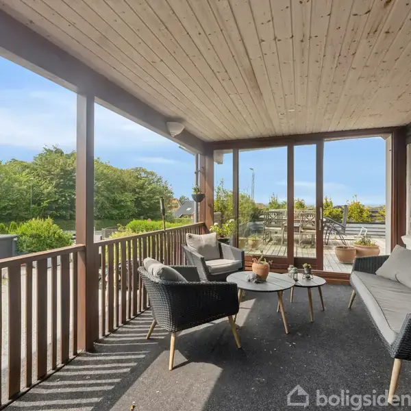 Veranda med komfortable møbler, herunder en sofa og to stole omkring et lavt bord. Beliggende i en have omkranset af grønne buske og træer, med klar blå himmel i baggrunden.