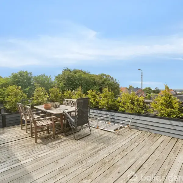 Træbord og stole står på en træterrasse, omgivet af frodig beplantning og med en udsigt over grønne områder og himmel. Flere potteplanter er placeret langs rækværket.