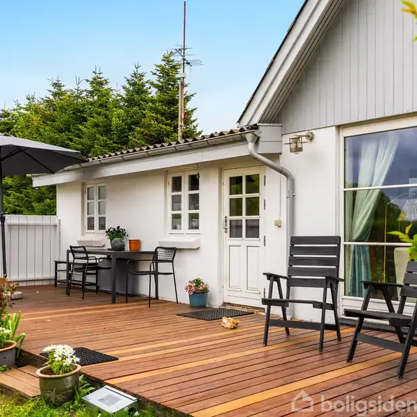 Hus med hvid facade, terrasse møbleret med to sorte stole og parasol. Blomsterkrukker og grønne planter omkranser terrassen. Lille bord ved siden af døren; træer i baggrunden.