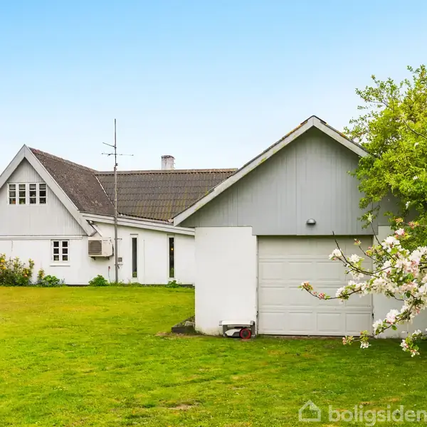 En hvid villa med mørkt tag står stille i en grøn have omkranset af blomstrende træer. Garagen er en integreret del af huset.