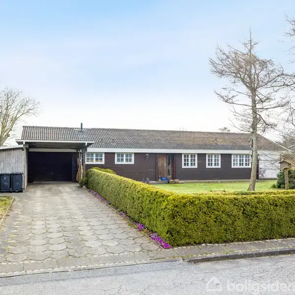 En lav, brun murstensvilla med garagedør på venstre side, flisebelagt indkørsel og omkranset af grønne hække i et villakvarter. Der står tre skraldespande ved garagen.