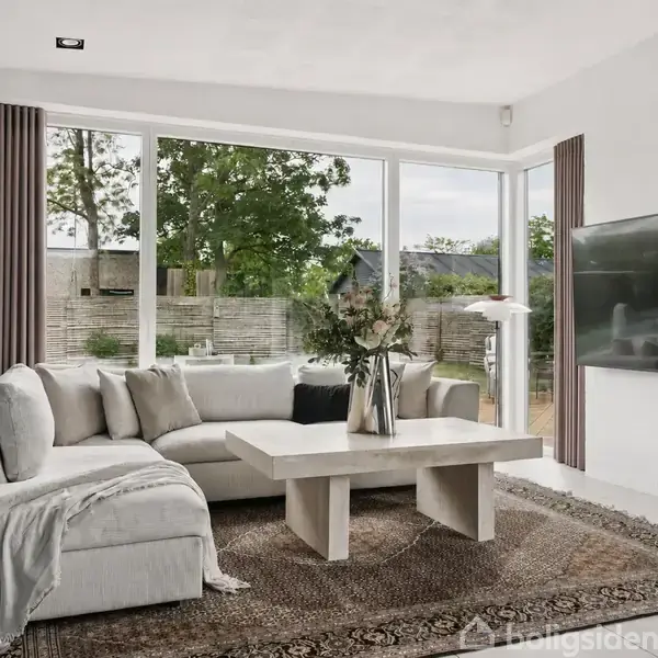 Stuen indeholder en grå sofa med tæppe, placeret omkring et moderne sofabord med blomster. Store vinduer viser en have bagved. En fladskærm hænger på væggen.