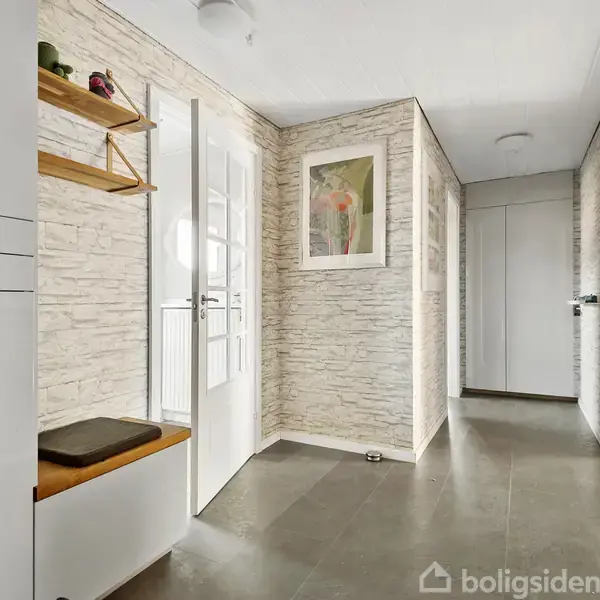 En lys gang med stenmønstret tapet og grå fliser. En hvid dør med glasindsats ses i midten. Venstre væg har hylder og højre væg viser indgang til et rum. En plakat hænger på væggen.