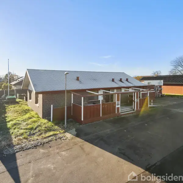 En rød murstensbygning står stille ved en vej, omgivet af grus og græs, med en klar blå himmel i baggrunden.