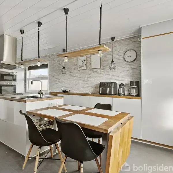 Et moderne køkken har et spisebord med to sorte stole, omgivet af skabe og hvidevarer. Loftslamper hænger over bordet. En vægur og køkkenmaskiner er på en bageste bordplade.