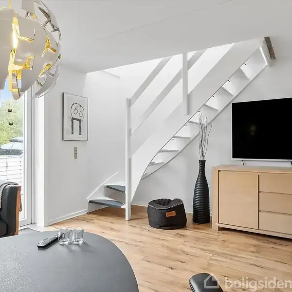 Lænestol med puder står ved runde bord i moderne stue; trappe leder opad ved siden af rektangulært TV på skab. Store vinduer giver lys. Tæppe og kunst på væggen.