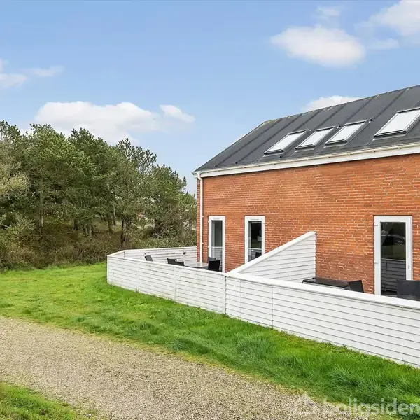 Et murstenshus med sorte tagvinduer står ved siden af en have med græsplæne. En hvid træterrasse omgiver huset. I baggrunden er der træer og en klar blå himmel.