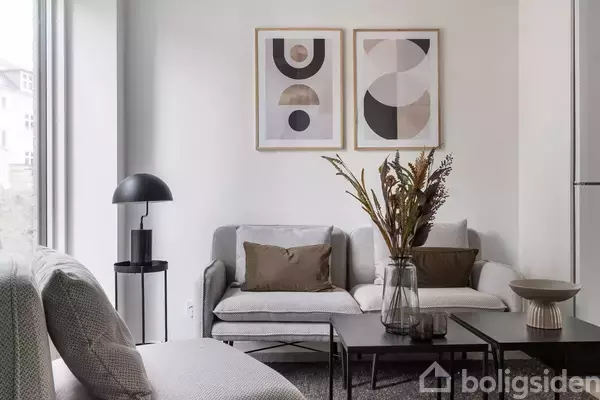 Sofa med brune puder står ved siden af sort bordlampe. To minimalistiske kunstværker hænger på væggen. En vase med tørrede blomster står på et sofabord i et lyst rum.