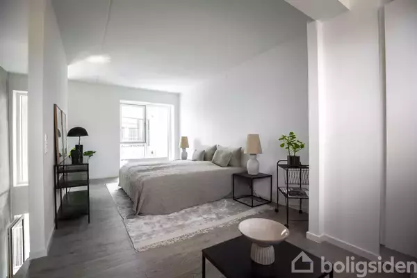 Soveværelse med dobbeltseng dækket med grå sengetøj, flankeret af natborde med lamper og planter. Store vinduer bringer naturligt lys ind, lyse vægge og trægulv i moderne design.