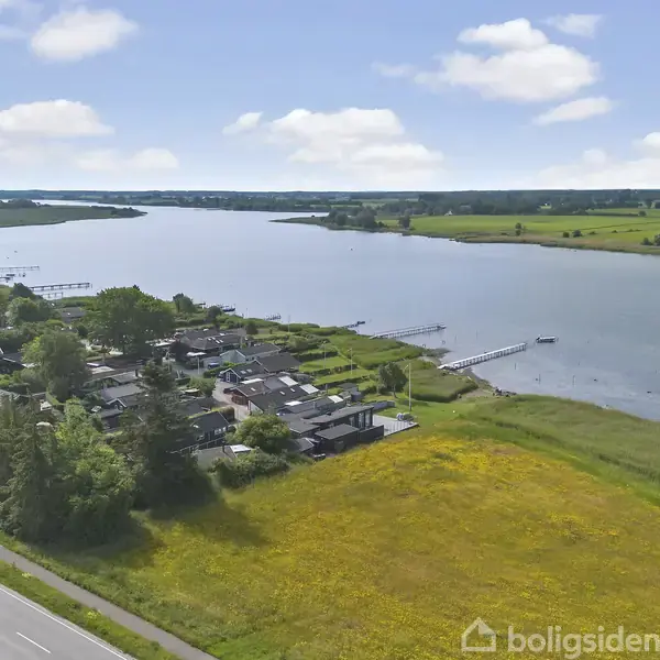 Boliger ved en flod strækker sig langs kysten, omgivet af frodige marker. En vej løber parallelt med floden, og en blå himmel med få skyer danner baggrunden.
