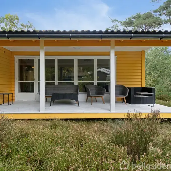 Et gulmalet sommerhus står stille med en veranda, hvor der er placeret sorte havemøbler. Omkring huset ses grøn natur og høje træer under en klar blå himmel.