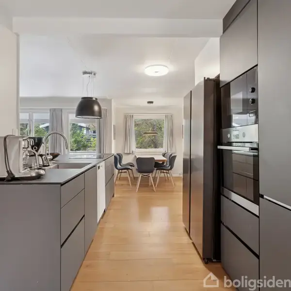Et moderne køkken med grå skabe og stålhovede apparater, som inkluderer en indbygget ovn og vask, åbner op mod en spisestue med et bord og stole, omgivet af vinduer.