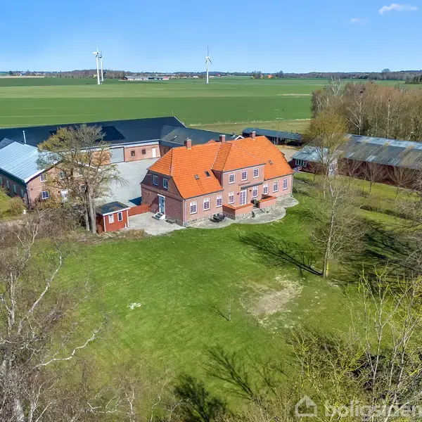 En stor murstensgård med rødt tag står i en grøn have. Bagved ses flere gårdbygninger. Omgivelserne er åbne marker med vindmøller i baggrunden under en klar blå himmel.