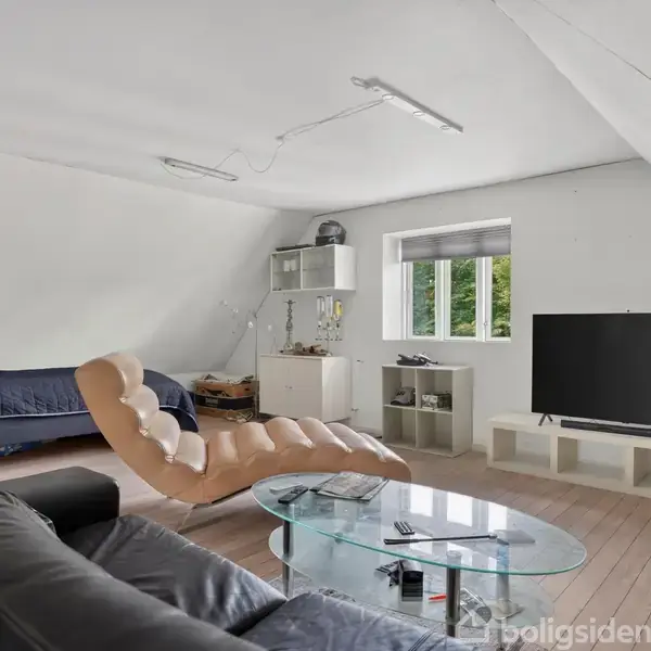 Et møbleret værelse med en sort lædersofa, glasbord og stor fladskærm. En seng er til venstre, og kasser står ved siden af en tv-bænk. Væggene er hvide.