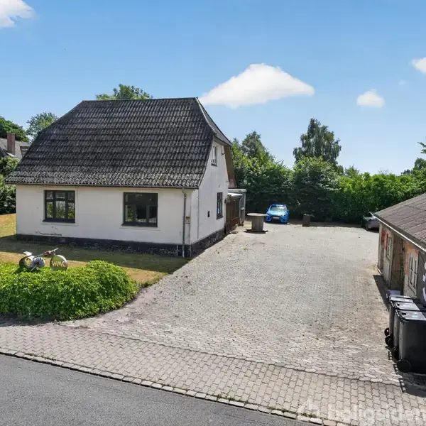 En hvid etplanshus med sort tag står på en brolagt gårdsplads. En blå bil er parkeret ved siden af huset, omgivet af grønne buske og træer.