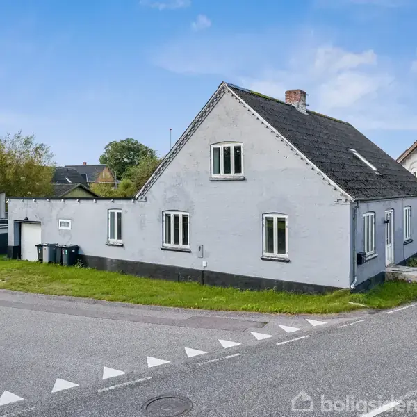 Gråmalet hus med sort tag og hvide vinduer står på hjørnet af to asfaltveje. Omgivet af grønne buske og en anden bolig ses i baggrunden under en blå himmel.