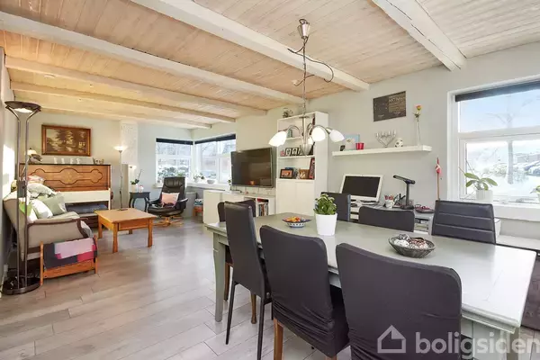 Stue med spisebord og seks stole i forgrunden. Sofahjørne med lænestol til venstre, TV på væggen. Store vinduer slipper lys ind. Loftet har synlige bjælker.