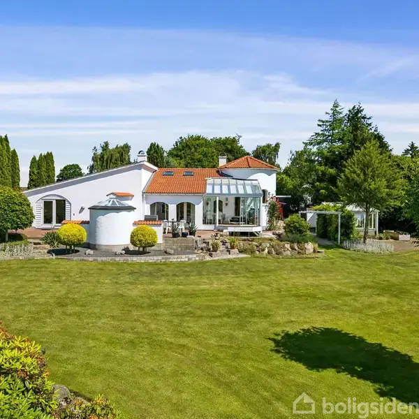 Hvid villa med orangerødt tag står omkranset af velplejede græsarealer og træer. En udestue er tilknyttet huset, og der ses store vinduer samt forskellige buske og planter omkring.