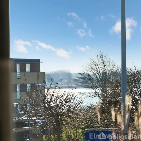 En vejskilt med teksten "Elmevej" står foran et træ og en bygning. I baggrunden skimtes en sø eller flod under en klar blå himmel med få skyer.