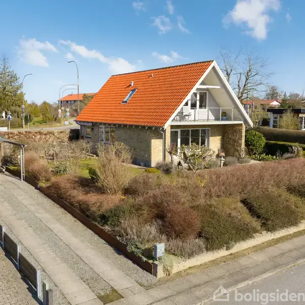 Et gult murstenshus med rødt tag står i en velholdt have omgivet af buske. En carport er placeret til venstre, og en vej løber foran huset under en blå himmel.