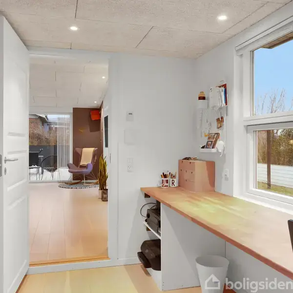 Et skrivebord med kontorartikler står ved et vindue, hvorfra der er udsigt til en have. En åben dør fører til en stue med lænestol og maleri.