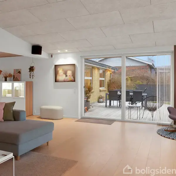 En moderne stue med en grå sofa står foran en stor glasdør, der fører ud til en terrasse med møbler. Rummet har kunst på væggene og elegant belysning.