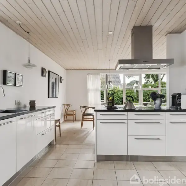 En moderne stue med en grå sofa og rød stol foran et stort vindue. Et TV hænger på væggen, og et tæppe dækker gulvet. Udenfor er der grønt.