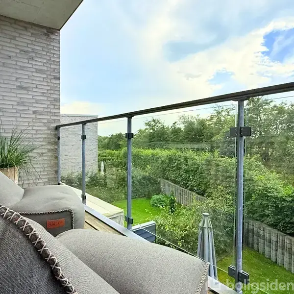 Balkon med beige sofaer står stille ved glasrækværk. En plante er placeret til venstre. Baggrunden viser en udsigt over en grøn have og blå himmel med skyer.