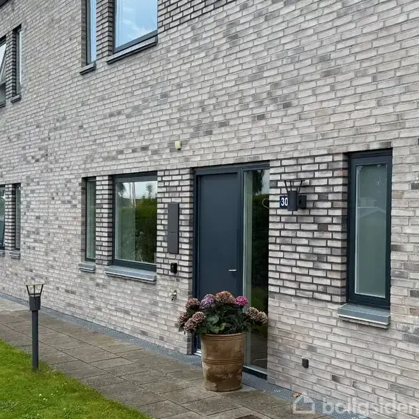 Rækkehus med grå mursten, sorte døre og flere vinduer. En blomsterkrukke står ved indgangen, og en plæne løber langs fortovet. Husnummer "30" er monteret ved døren.