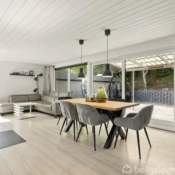 En moderne stue med spisebord og seks stole; store vinduer giver udsigt til terrasse og have. En brændeovn står ved siden af en lys sofa i hjørnet.