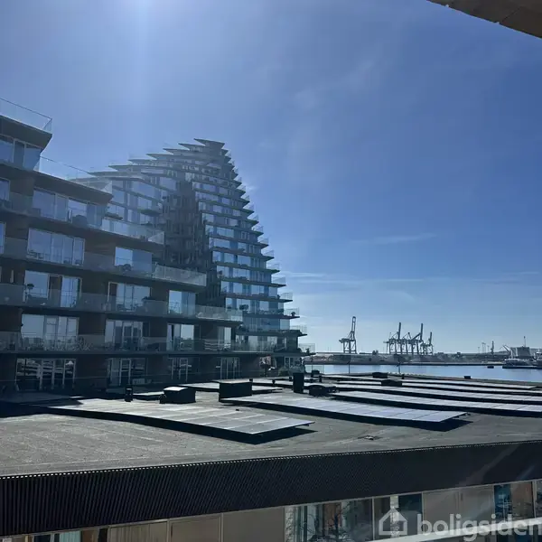 Et moderne boligkompleks med terrasse og glasfacade står ved havnen. Baggrunden viser kraner og vand under en klar blå himmel.