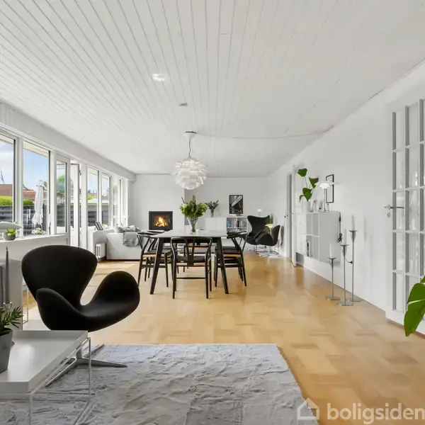 Stue med sorte møbler og grønne planter, bordet står midt i rummet, omgivet af lyse vægge og store vinduer. Pejs i baggrunden, som skaber en hyggelig atmosfære.