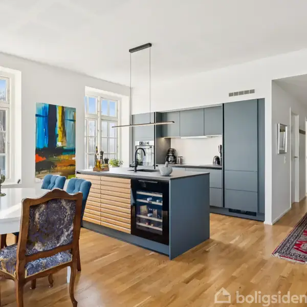 Et moderne køkken har et centralt køkkenbord med indbyggede apparater. Omkring er der en spisestue med træstole ved et rundt bord. Store vinduer giver lys, og en farverig maleri hænger på væggen.