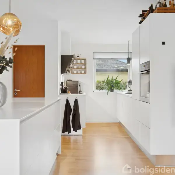 En moderne køkkenindretning med hvide skabe og træparketgulv. En glasvase med tørrede blomster står på bordet. En brun dør ses til venstre, med udsigt til en have gennem vinduet.