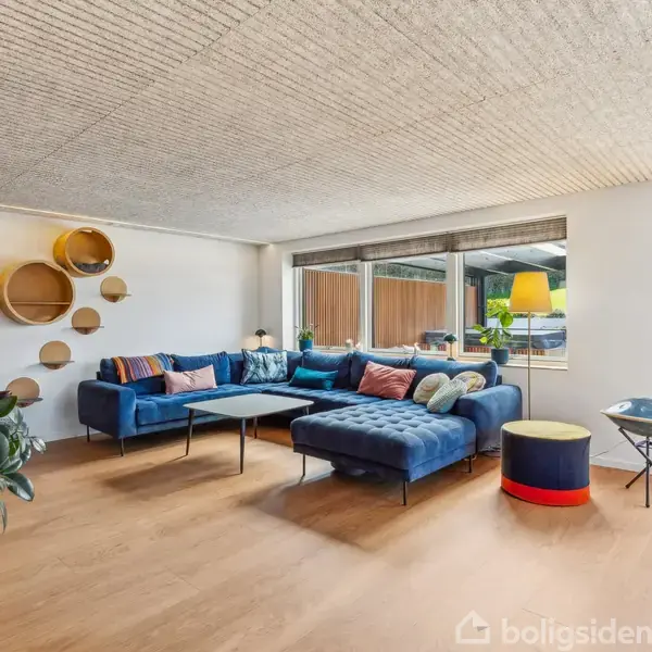 Blå hjørnesofa står i en lys stue med trægulv og stort vindue. Runde hylder på væggen og gulvlampe ved siden af sofaen. Grønne planter skaber en hyggelig atmosfære.