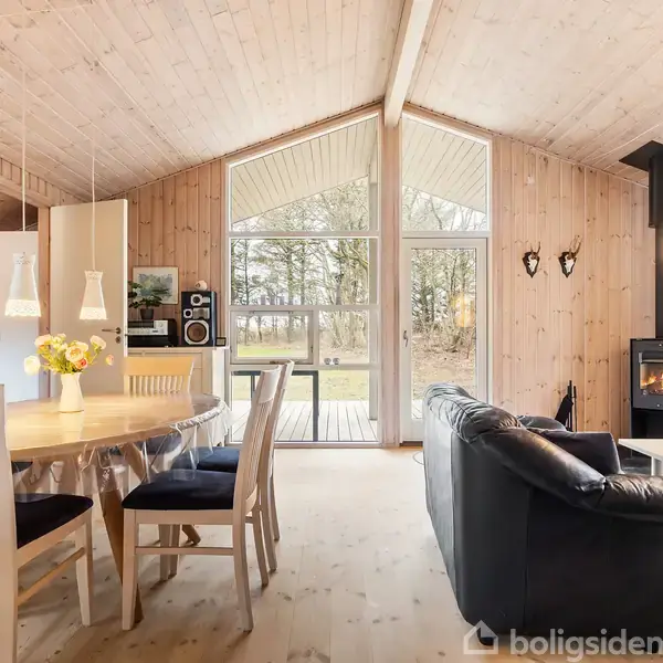 Spisestue med trævægge og -loft, spisebord med stole og blomster. Sofa ved siden af brændeovn. Store vinduer viser udsigt til have med træer. Afslappet atmosfære.