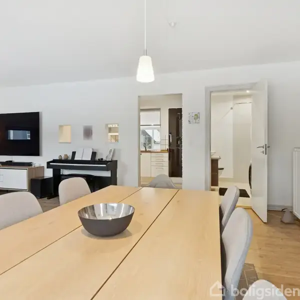 En lys spisestue med et langt spisebord omgivet af grå stole. En fladskærm hænger på væggen; rum indrettet med lyse trægulve og skandinavisk design i et hjemligt miljø.