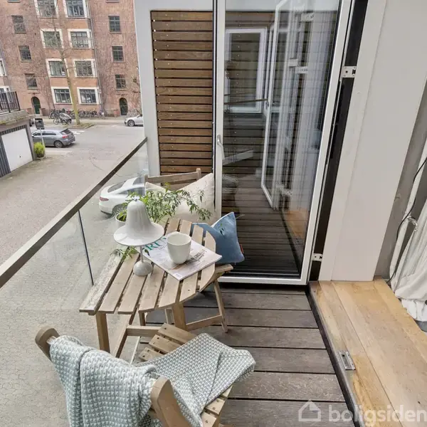 En lille altan med et rundt træbord, hvorpå der står en lampe og en kop. En stol med et tæppe befinder sig ved siden af. Bygning ses i baggrunden.