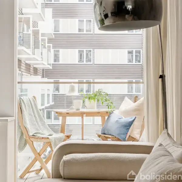 Stue med beige sofa og puder vender mod en balkon. Balkonbord med plante og krus, to træstole og tæppe i lejlighedskompleks.