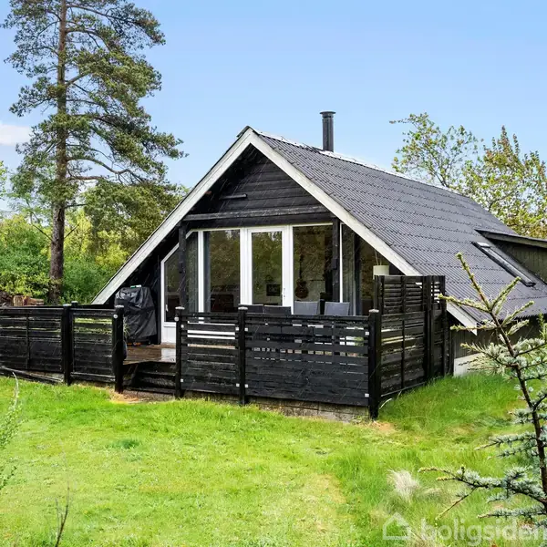 Et sortmalet træhus står stille i en frodig have med græs og træer, der omgiver strukturen. Huset har en terrasse med et hegn og store vinduer i gavlen.